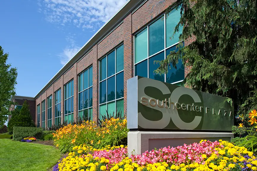 Exterior of a modern brick office building with large windows and landscaped flower beds. A sign in front reads "Southcenter Plaza" with large "SC" letters, surrounded by colorful flowers and greenery.